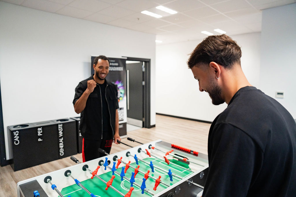 Two adult men wearing black casual clothing play table football in an office breakout room, one smiling and raising a fist in celebration during the game.