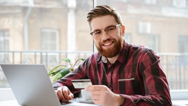 Man wearing glasses sitting in front of a computer with a credit card in hand