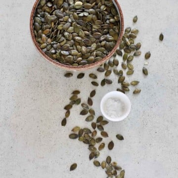 a bowl of pumpkin seeds on a grey surface beside a small bowl of sea salt