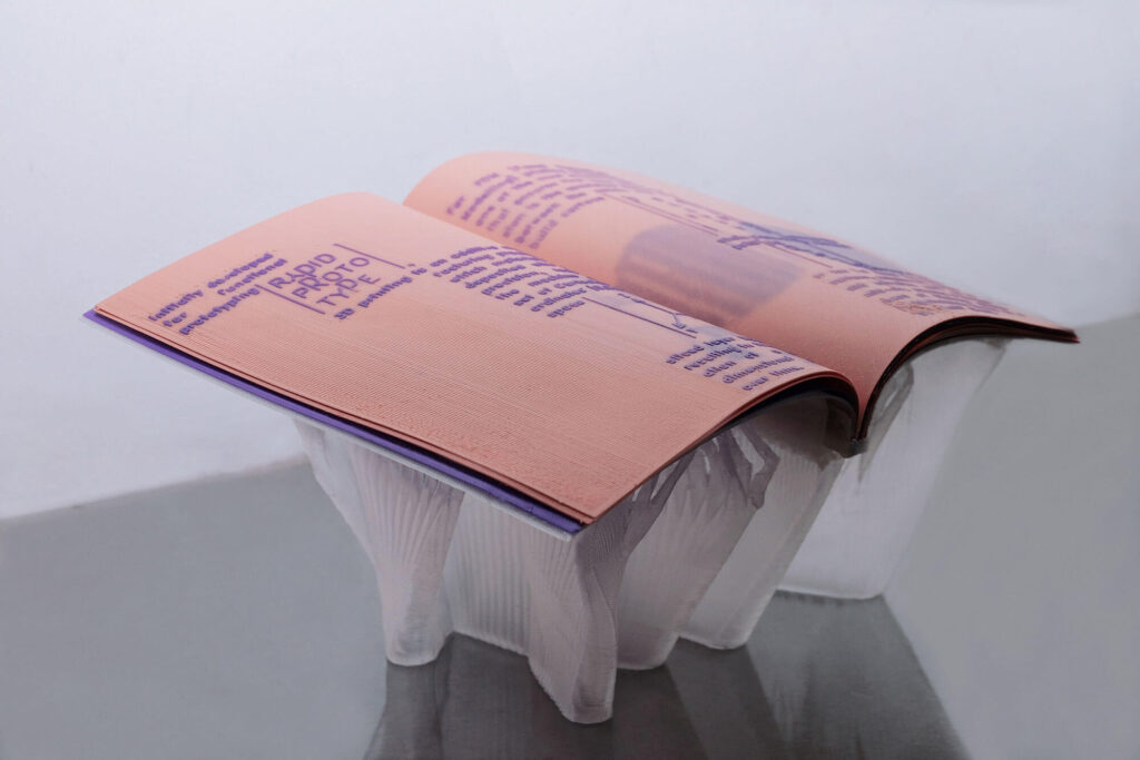 An open 3D printed book with pink pages and purple text rests atop a translucent, plastic container on a reflective surface, set against a plain, light background.
