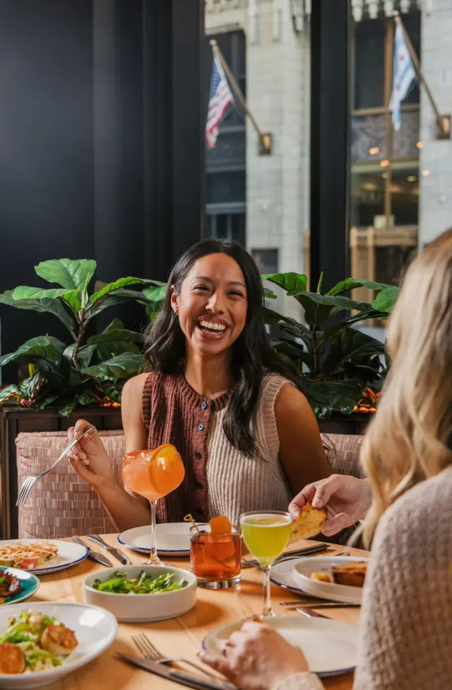 A joyful woman laughs while dining at Kindling inside the Willis Tower. The table holds wood-fired shared plates like scallops and diverse craft cocktails, including an orange spritz and an Old Fashioned. Through the massive window, the American and Chicago flags are visible amidst classic Loop architecture.