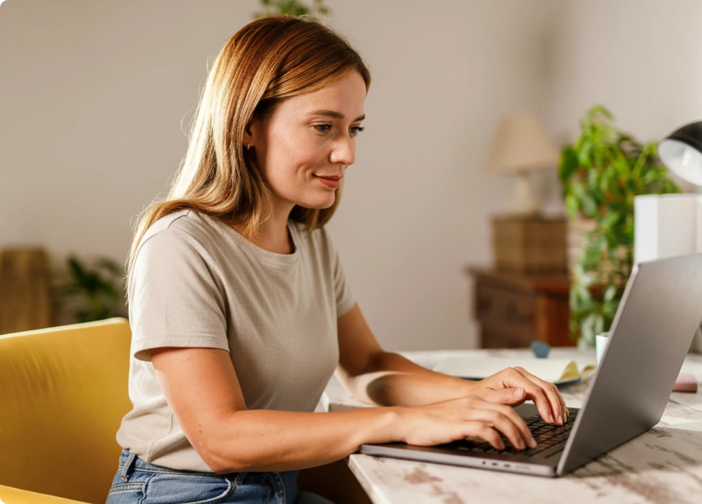 A woman with light brown hair sits at a table, typing on a laptop. She is wearing a beige t-shirt and appears focused. There are plants and home office items in the background.