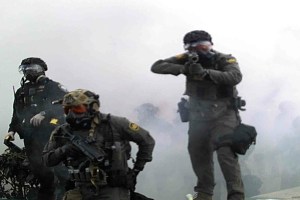 A federal agent in a uniform aims a weapon at the camera. Two other agents wearing gas masks and uniforms are to the agent’s right. There is smoke and outlines of more people behind them.