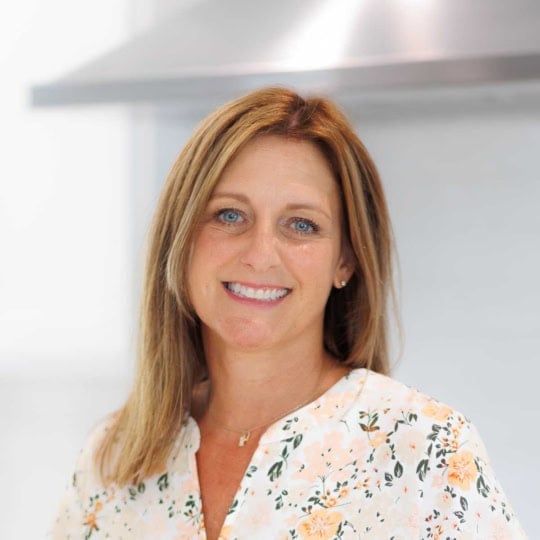 Woman with straight, light brown hair smiling, wearing a white floral blouse, standing indoors.