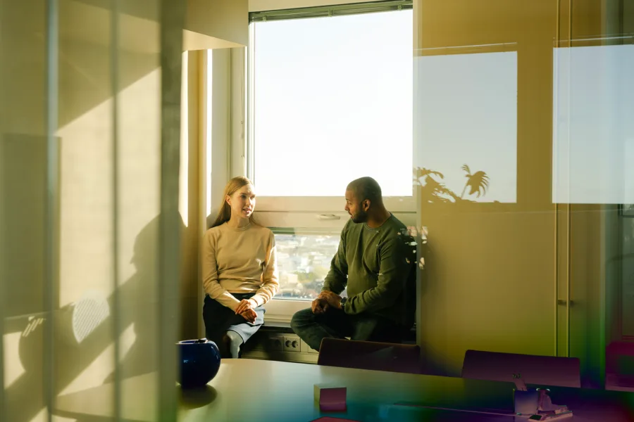 Two people sitting next to a window, in a brightly lit office.