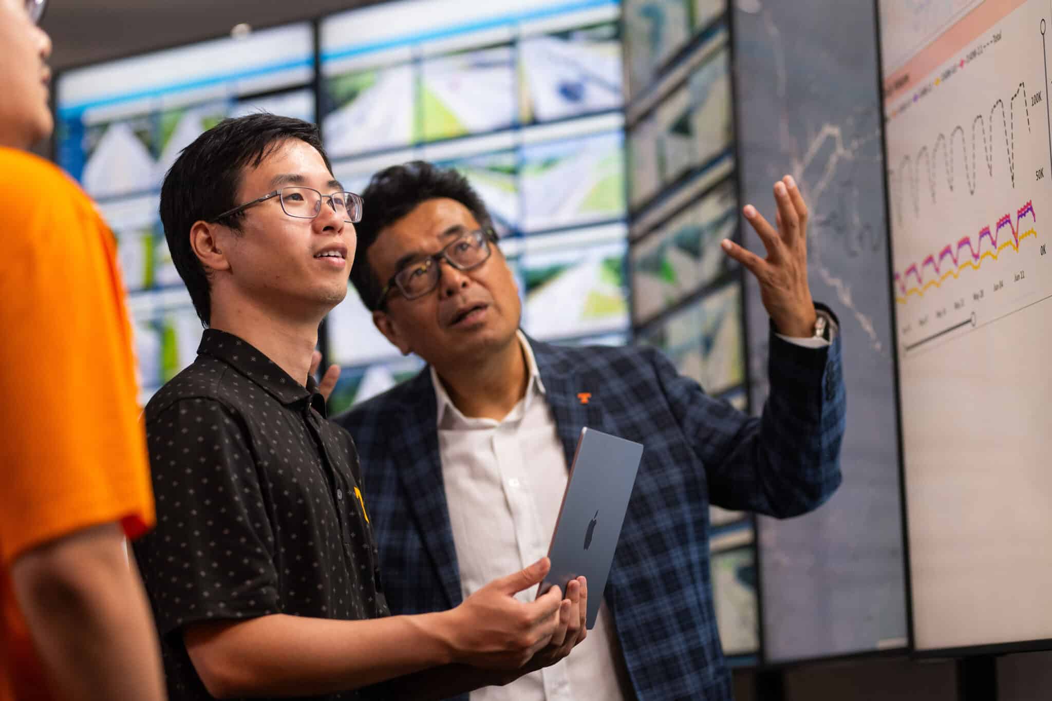 A faculty member discusses something with a student while gesturing at a giant video screen